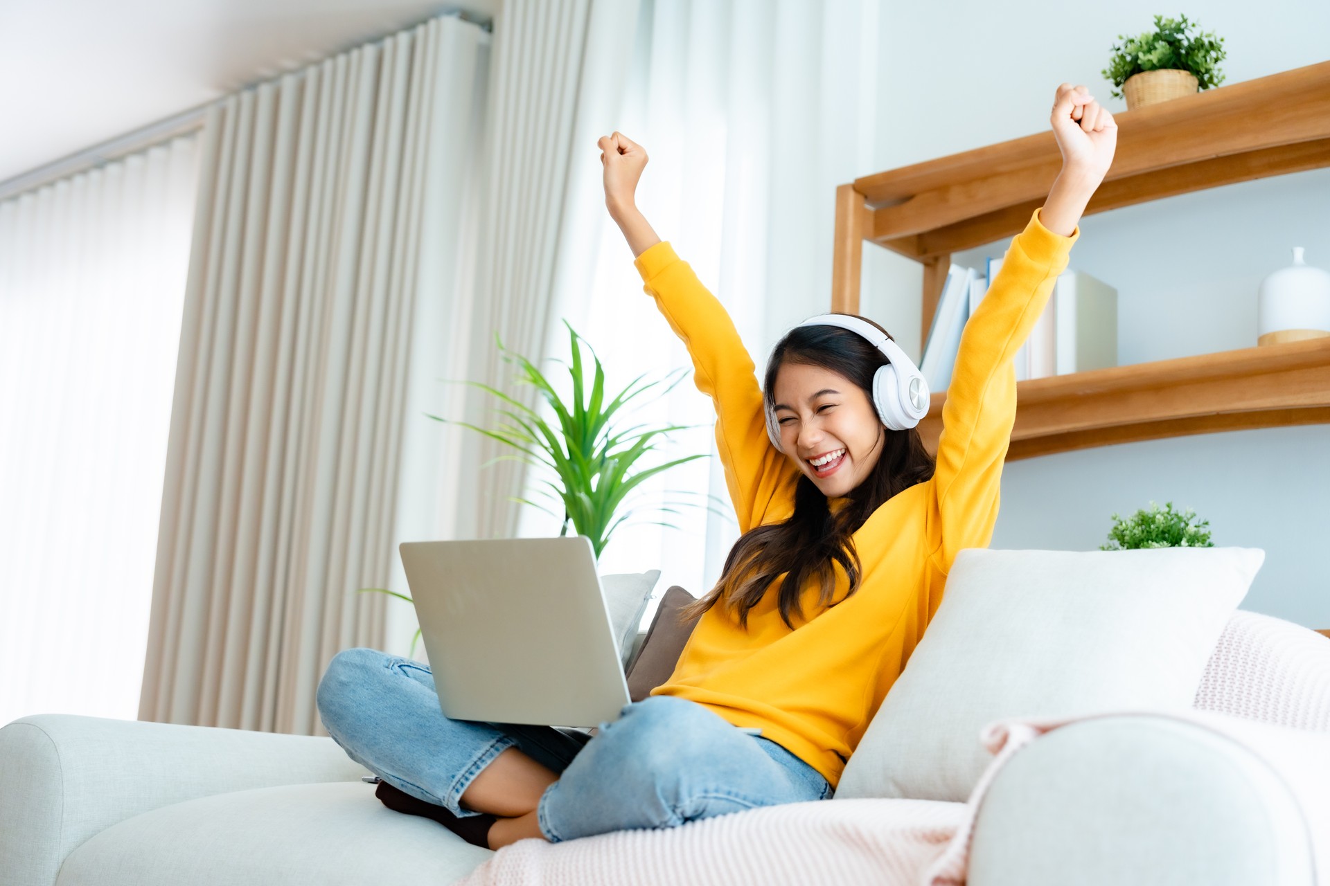 Young happy lucky woman student feeling excited winning, using computer laptop and sitting on sofa, adorable Asian female receiving great news on notebook, getting new job celebrating success